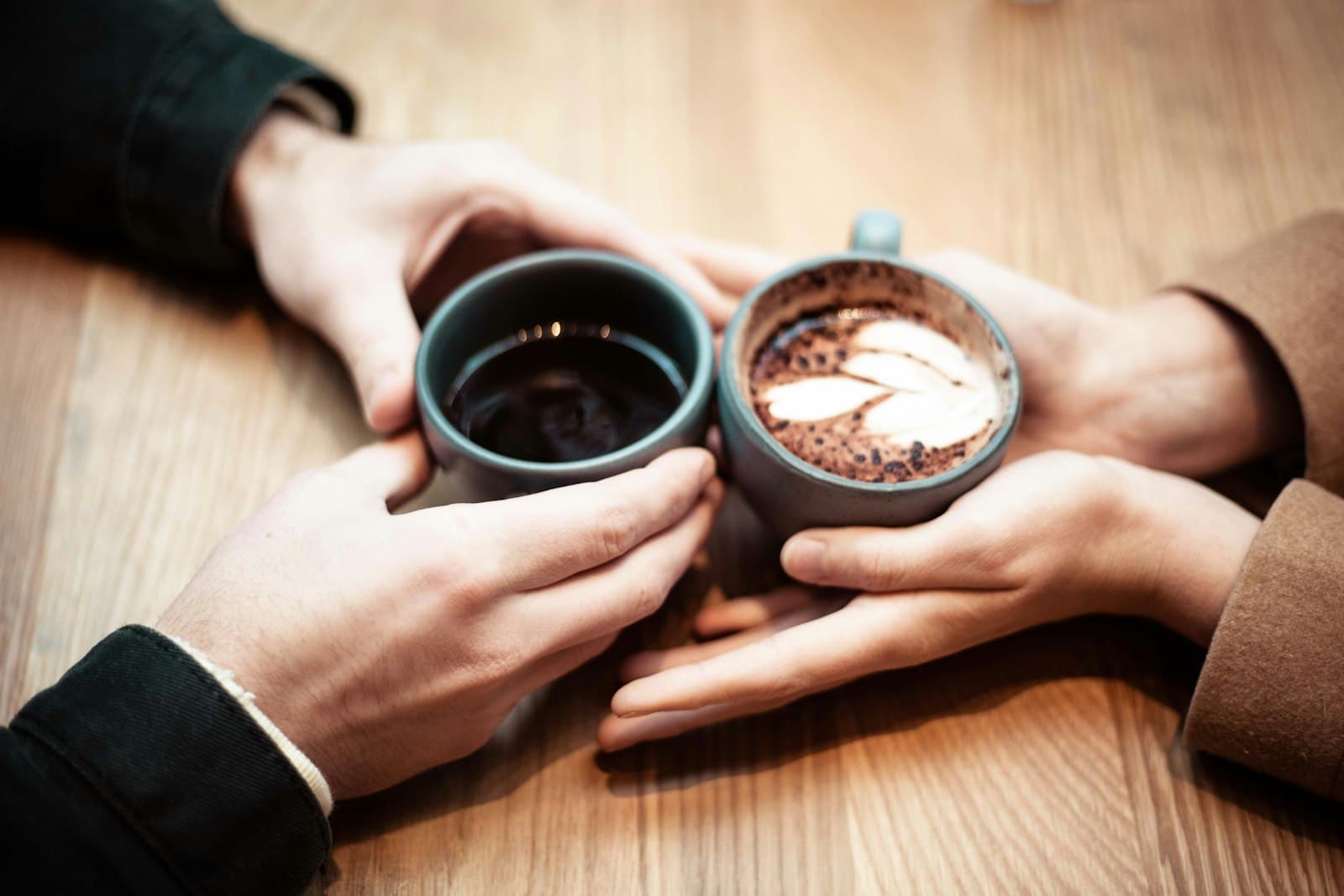 Twee mensen houden samen hun koffiemokken vast aan een houten tafel, een warm beeld van een gedeeld moment