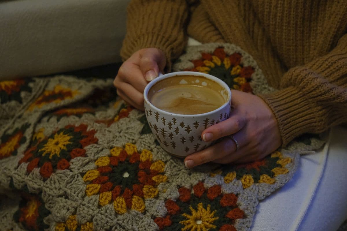 Handen houden een warme kop koffie vast boven een kleurrijke gehaakte deken tijdens een knusse avond thuis in het najaar