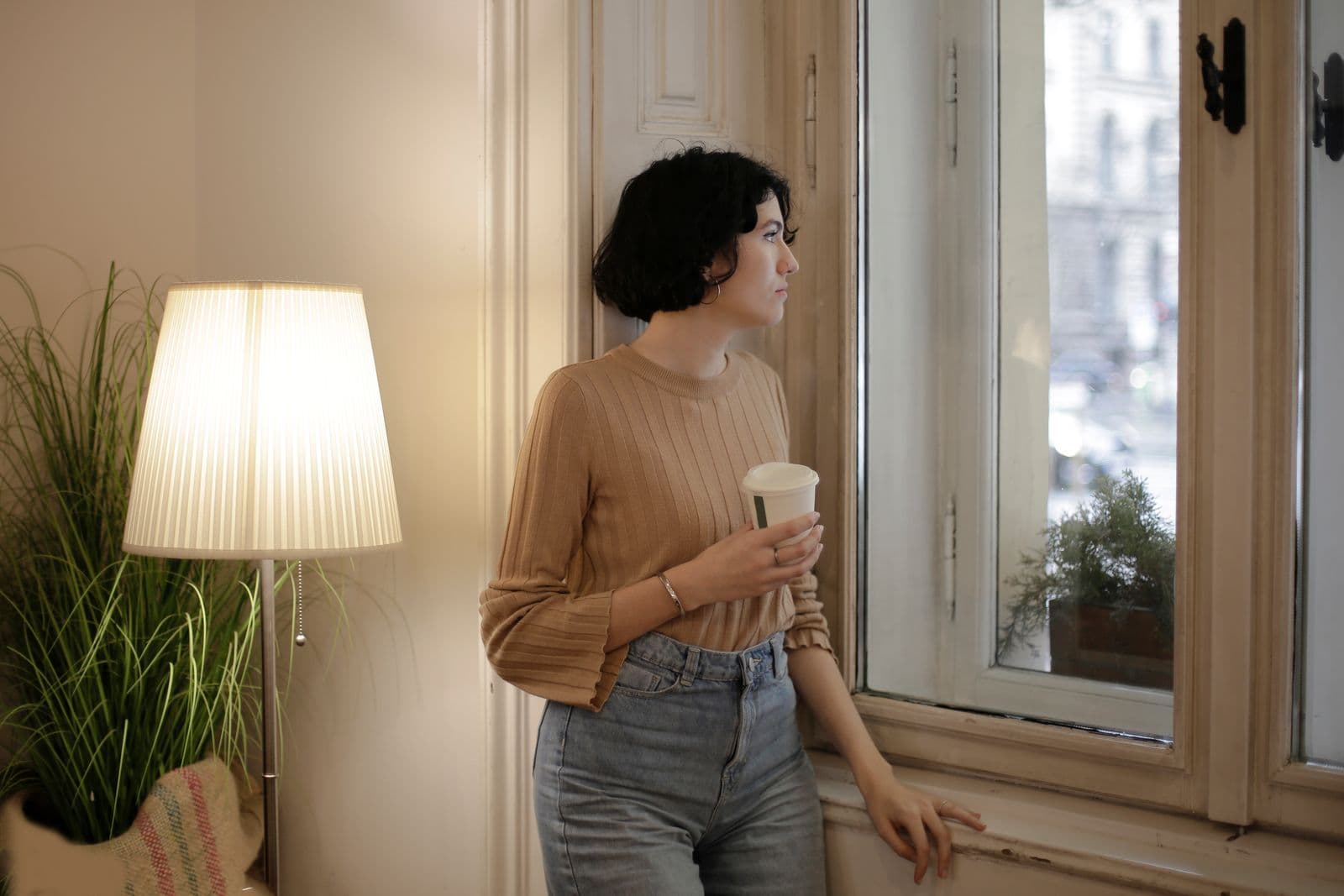 Een vrouw staat met een warme drank bij het raam naar buiten te kijken, een rustig moment waarop je aan iemand denkt die ver weg is