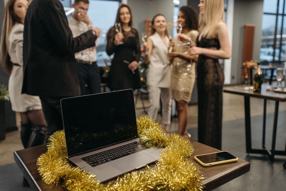 Een laptop met gouden slingers op een kantoortafel terwijl collega's op de achtergrond met glazen proosten op het einde van het werkjaar