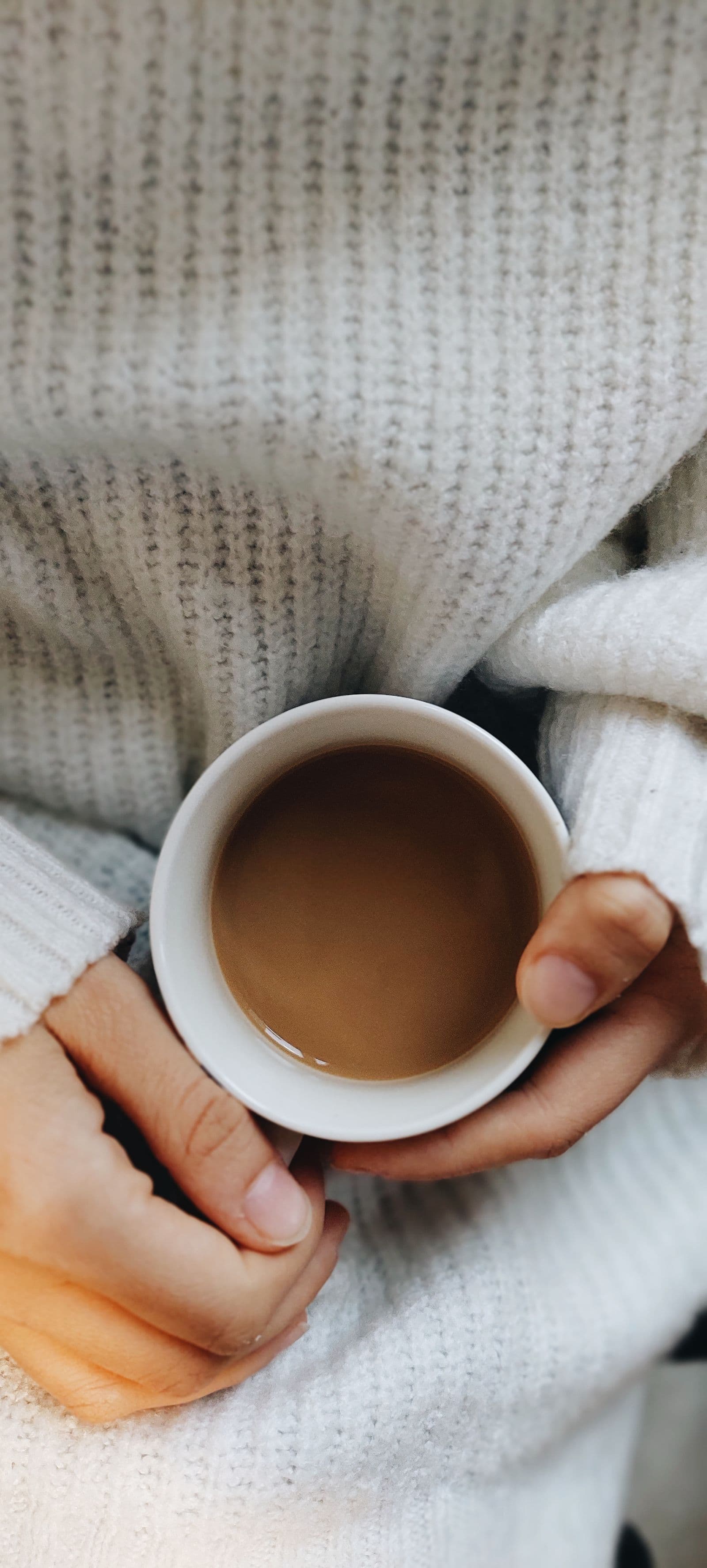 Twee handen in een zachte trui houden een warme mok koffie vast, een rustig en huiselijk beeld bij een koffiemoment met een koekje