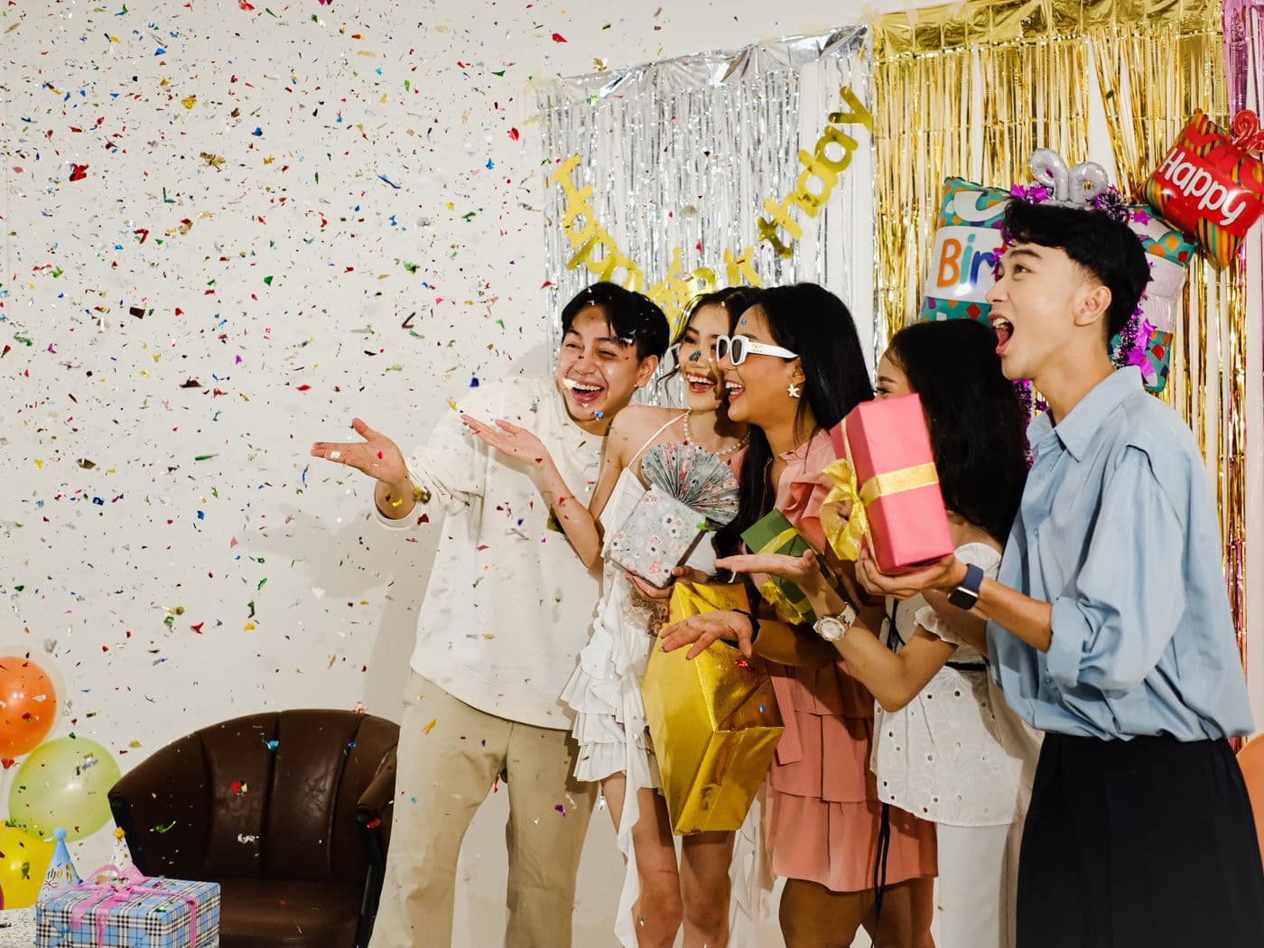 Een vrolijke groep vrienden viert lachend een verjaardag met confetti en cadeaus in de hand onder een slinger, een feestelijk beeld bij koekjes in plaats van taart