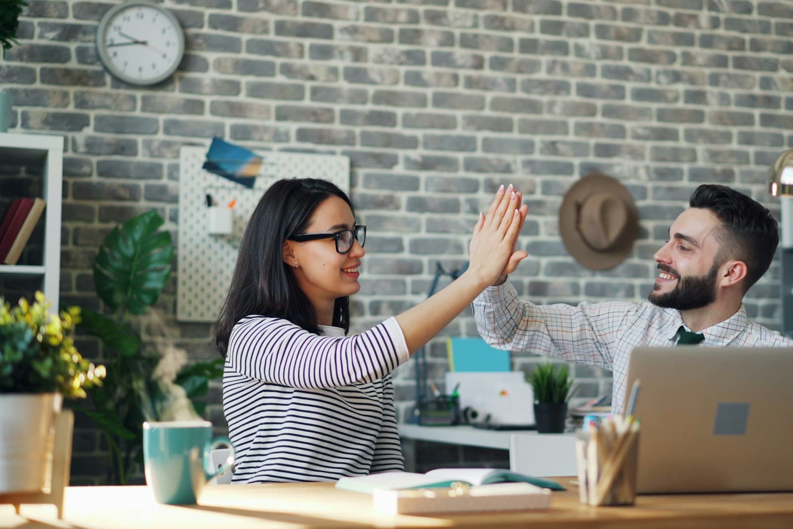 Twee collega's geven elkaar een high five aan een bureau met een laptop, een beeld van waardering en teamgevoel op de werkvloer