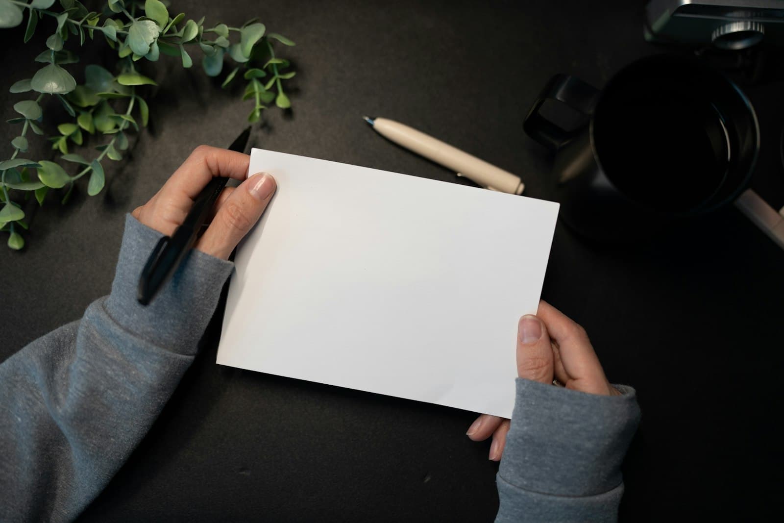 Handen houden een blanco kaartje met een pen boven een bureau met een plant en een kop koffie, klaar om een bedankje te schrijven