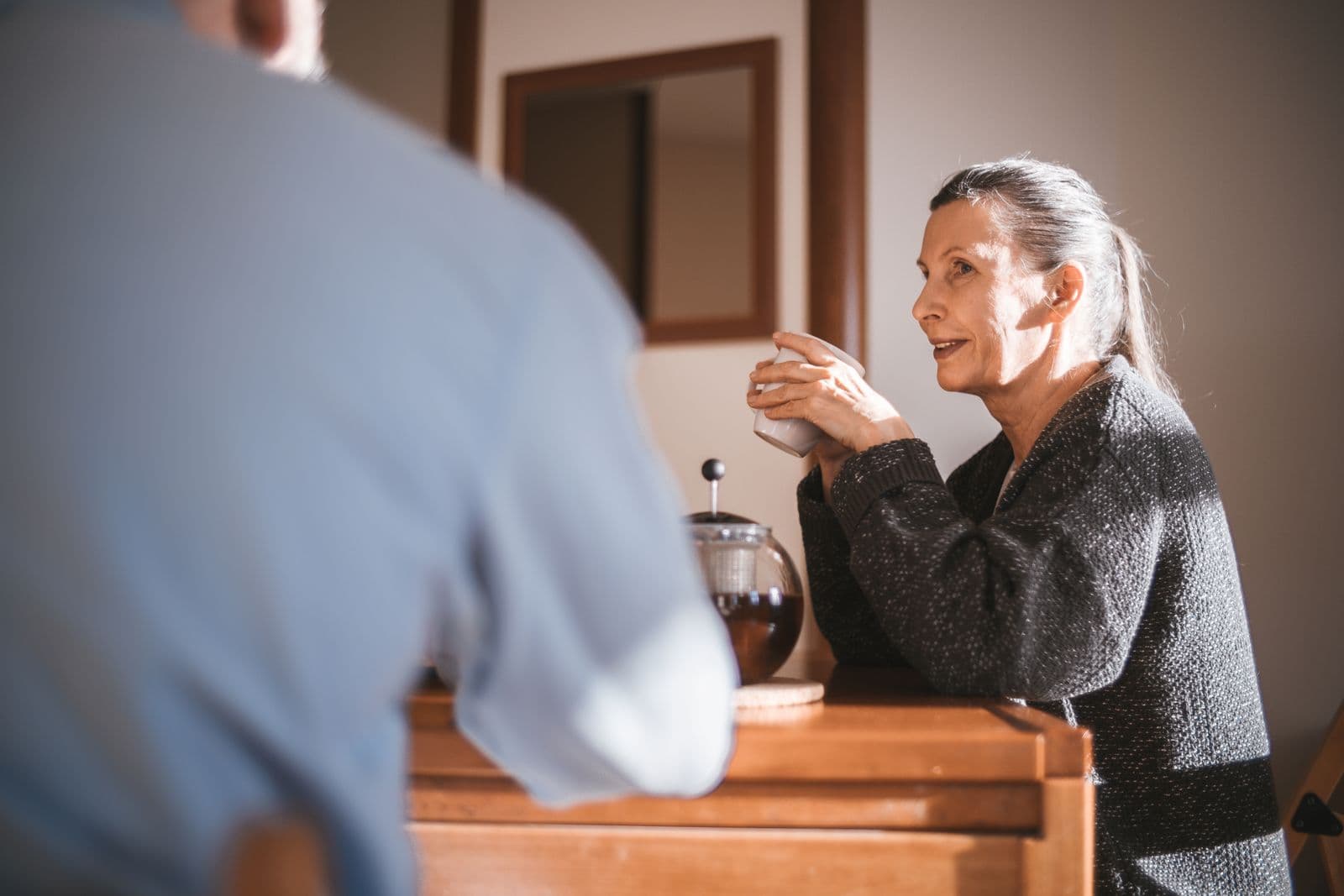Een oudere vrouw geniet thuis van een warme drank bij het raam, een rustig beeld bij een doos koekjes bij de koffie