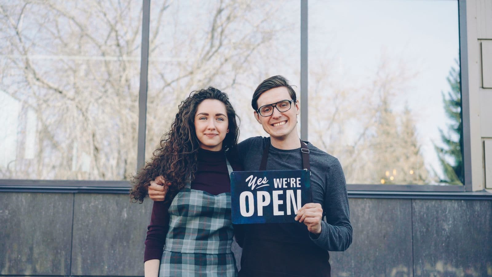 Twee trotse ondernemers staan voor hun nieuwe zaak met een bordje waarop we are open staat, een warm beeld bij de opening van een bedrijf