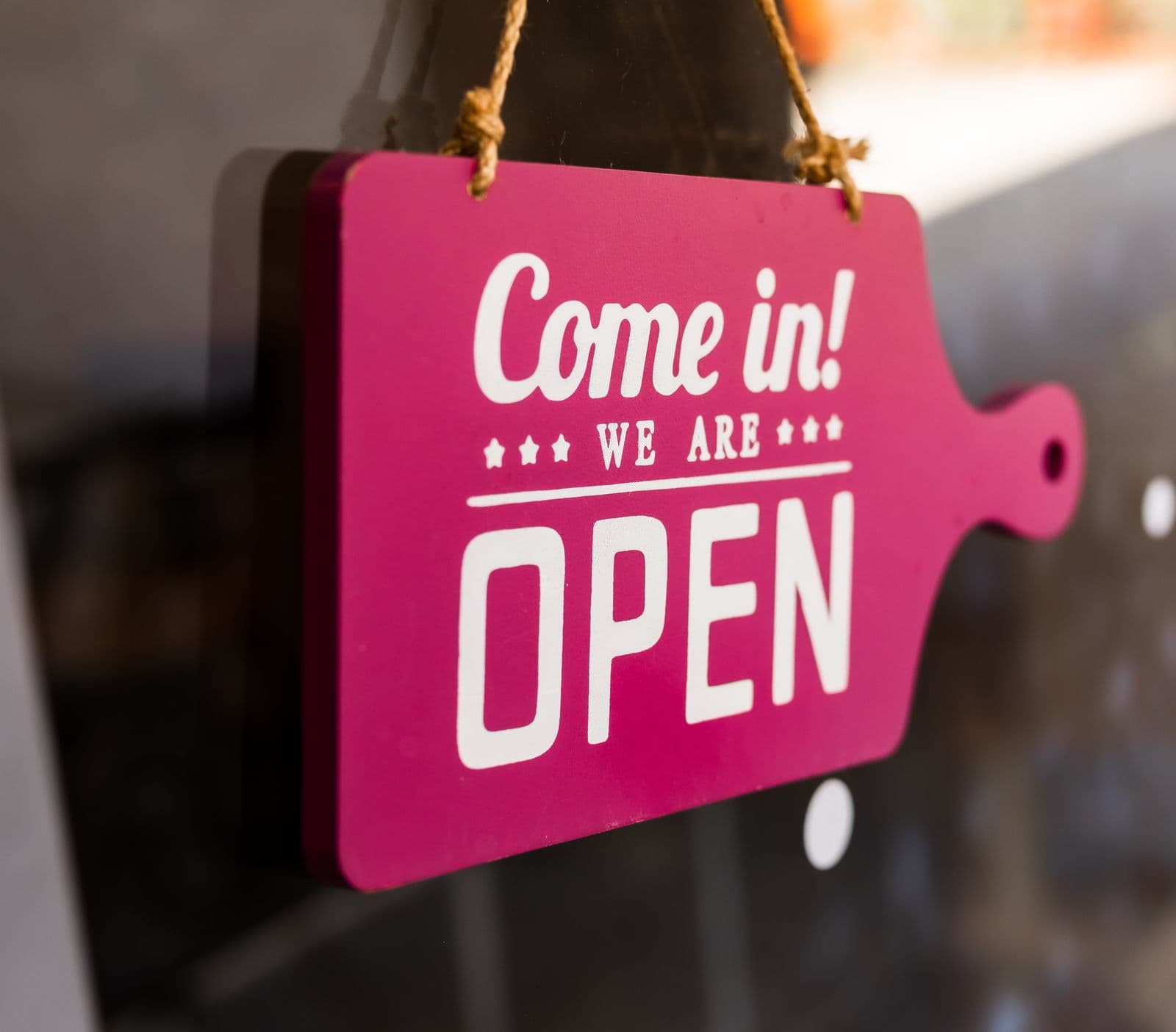 Een vrolijk roze bordje met de tekst come in we are open hangt in een winkelruit, een uitnodigend beeld bij de opening van een zaak