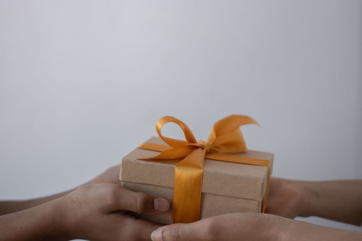 Twee handen overhandigen een in kraftpapier verpakt cadeaudoosje met een oranje lint als meeneemcadeau