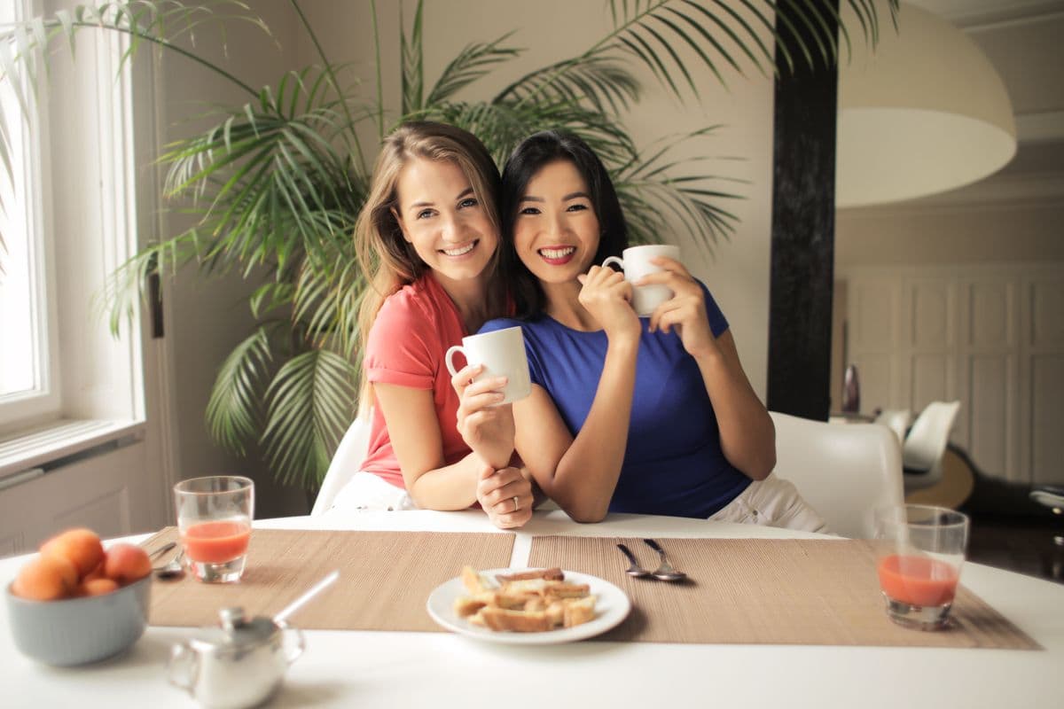 Twee vriendinnen zitten lachend samen aan een lichte tafel met een kop koffie in de hand tijdens een gezellig bezoek thuis