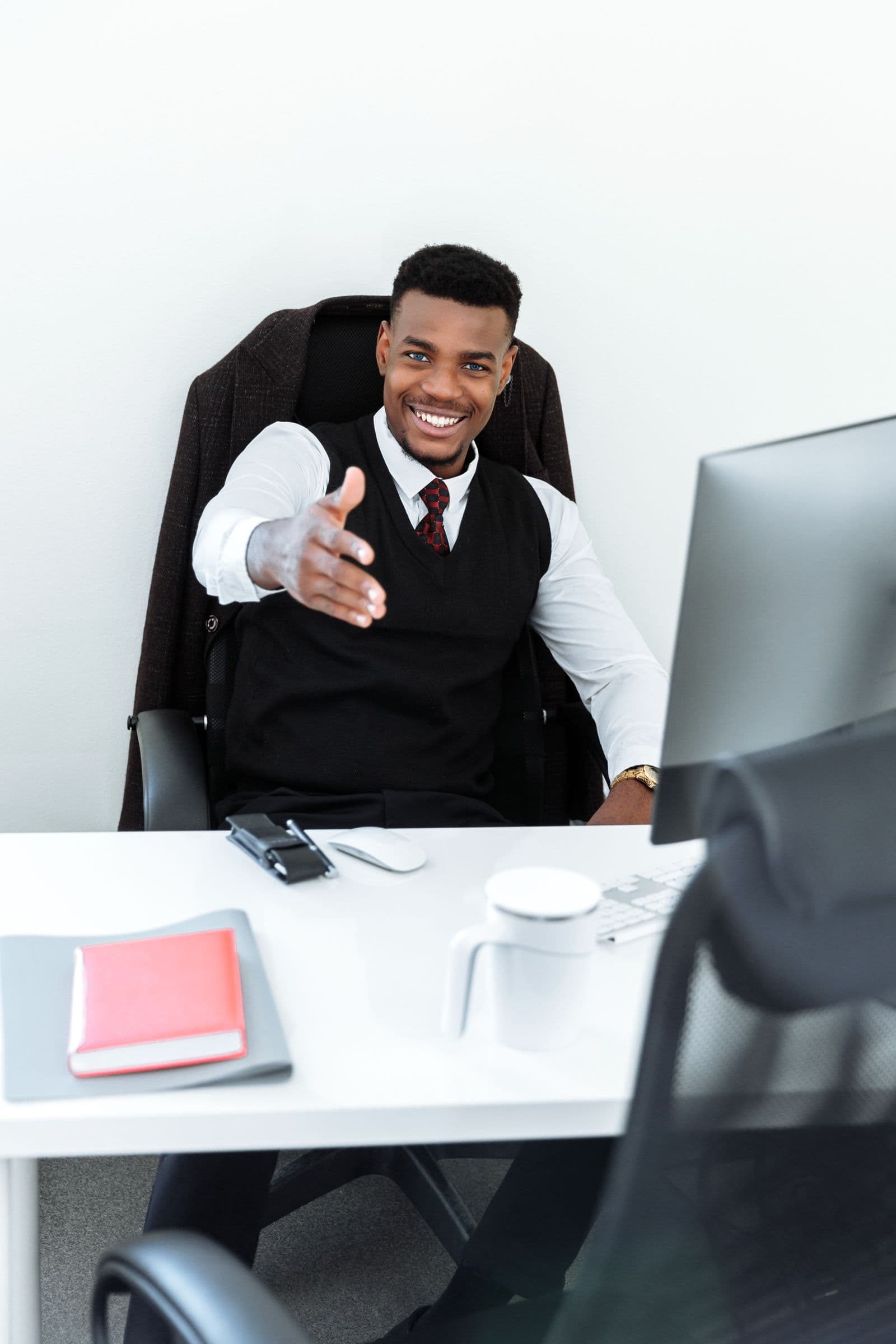 Een glimlachende medewerker aan een bureau reikt zijn hand uit om iemand te verwelkomen, een warm beeld bij een nieuwe collega die welkom wordt geheten
