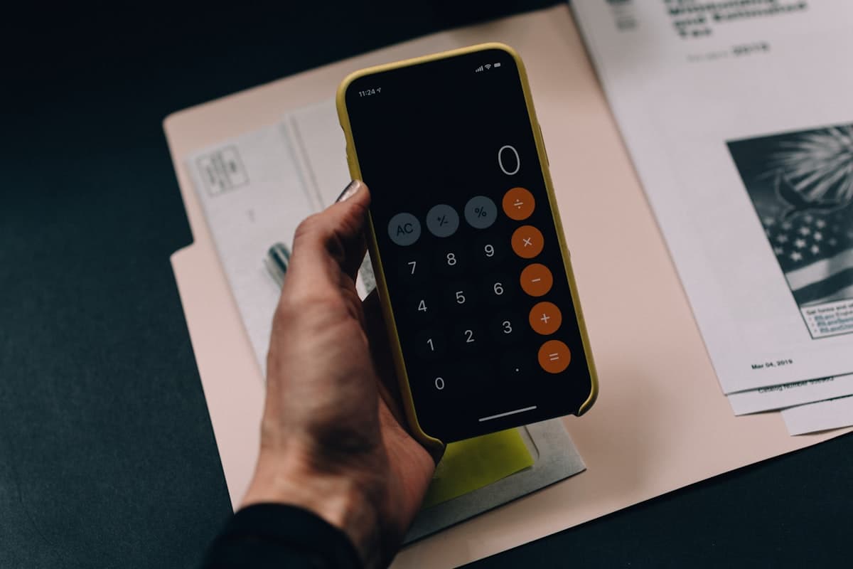 Een hand rekent met een rekenmachine boven een map met papieren op een bureau, passend bij het regelen van een factuur voor een zakelijke koekjesbestelling