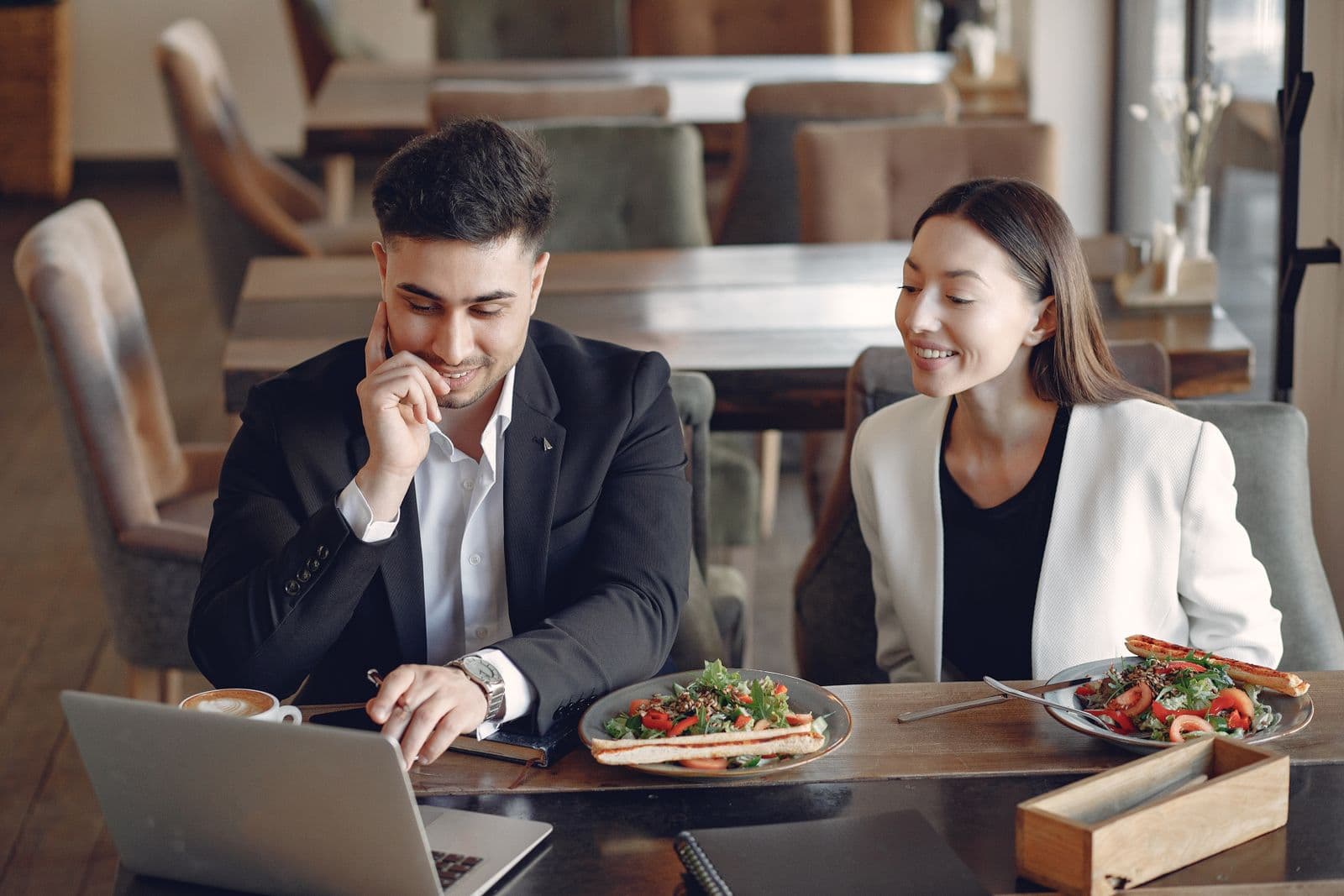 Een zakelijk duo lacht en overlegt aan een tafel met een laptop en een verzorgde lunch, passend bij koekjes bestellen voor een zakelijke lunch